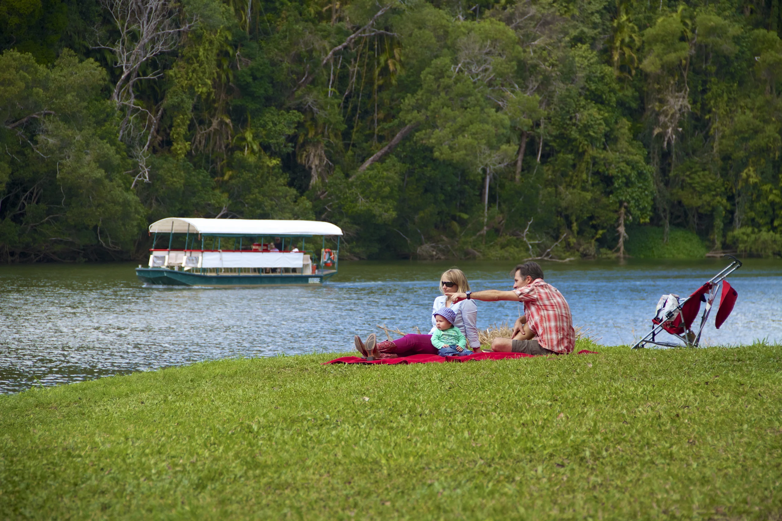 Two people sitting on the green grass at the edge of a river. A white boat can be seen in the water. 