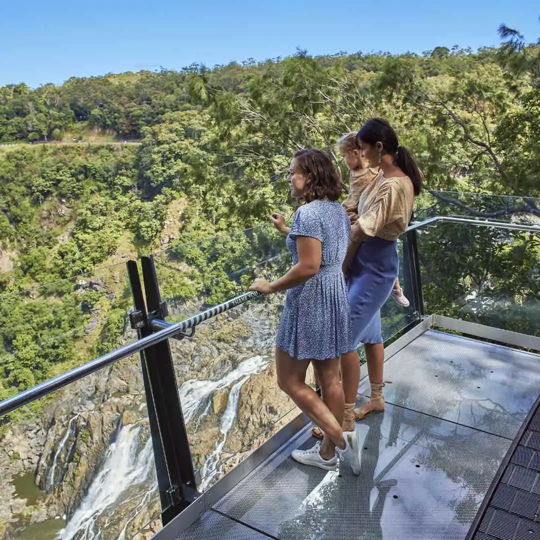 Two ladies and a small child stand on the glass section of the edge lookout looking up the Barron Gorge