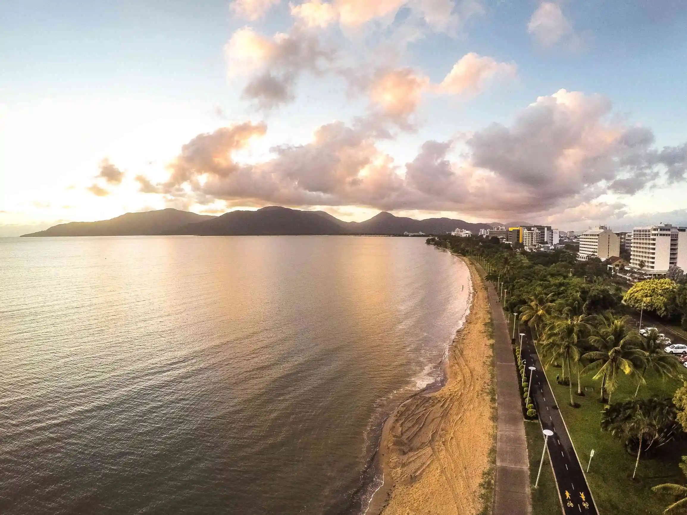 A view of cairns city edge fringed with palm trees and sand meeting the sea, mountains in the background.