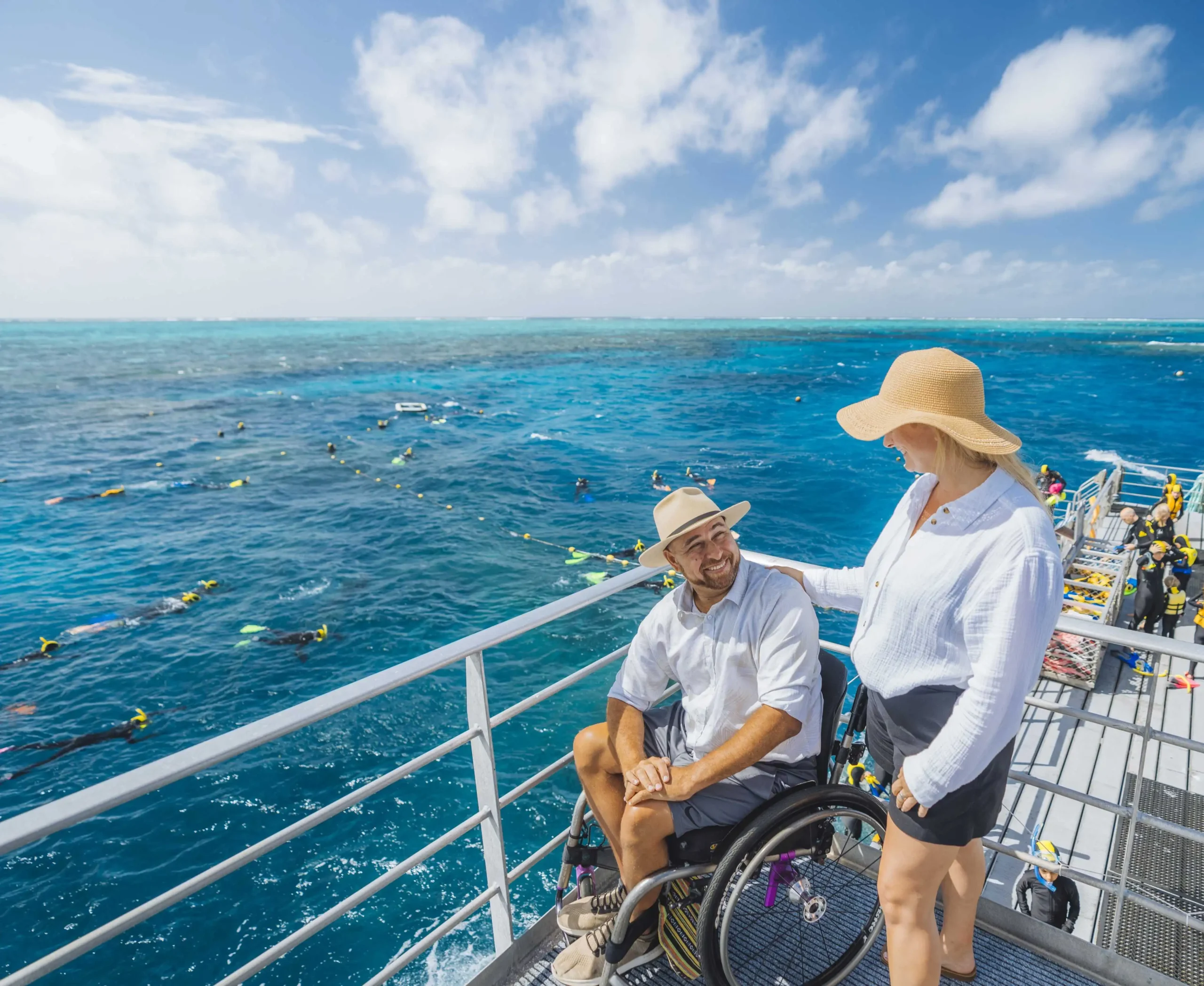A female wearing a sun hat and a male wheelchair user smiling on a cruise deck visitng the bright blue reef.