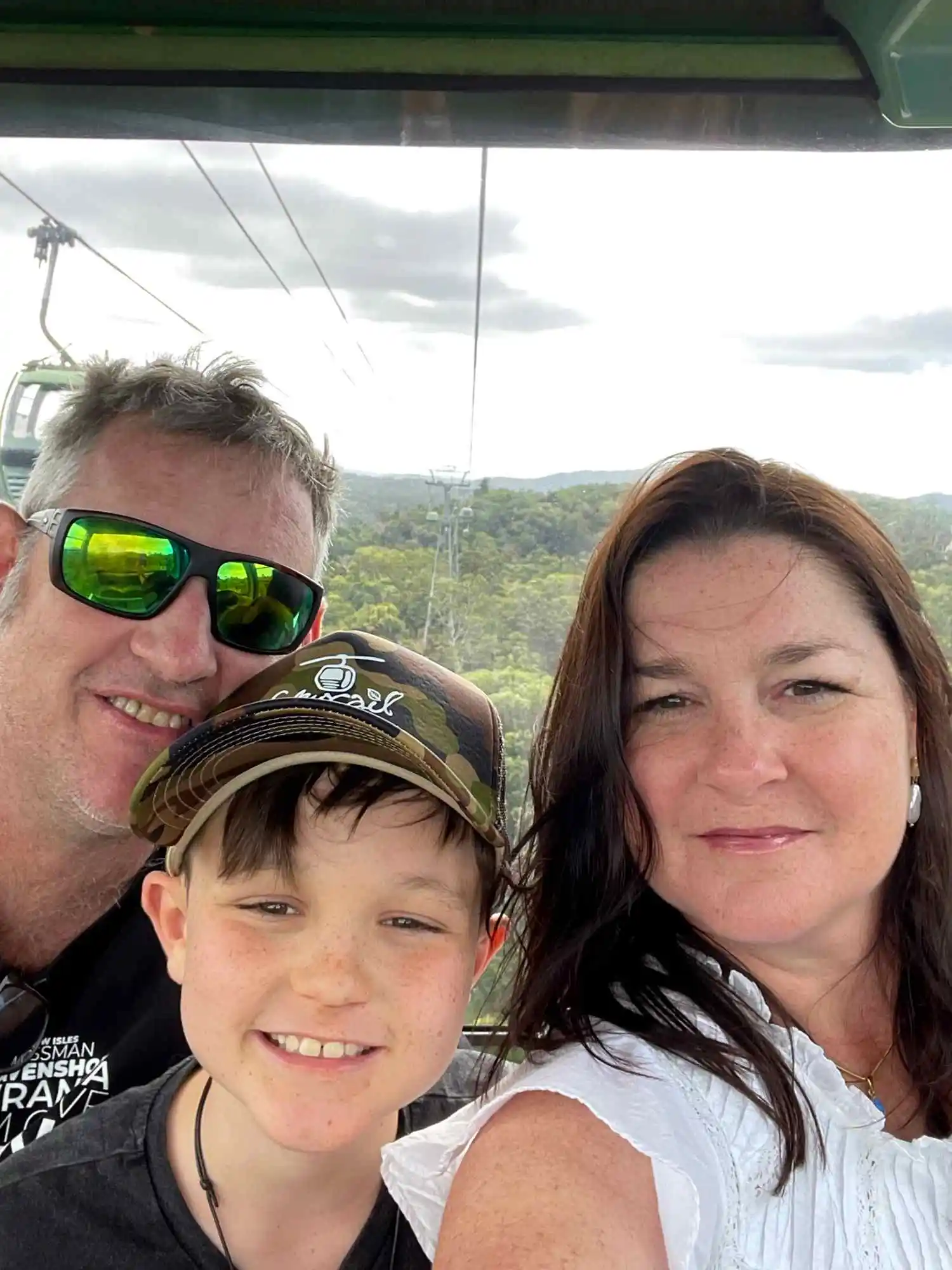 I love Cairns family on Skyrail A mum, dad and son smiling at the camera with a rainforest hill in the distance behind them.
