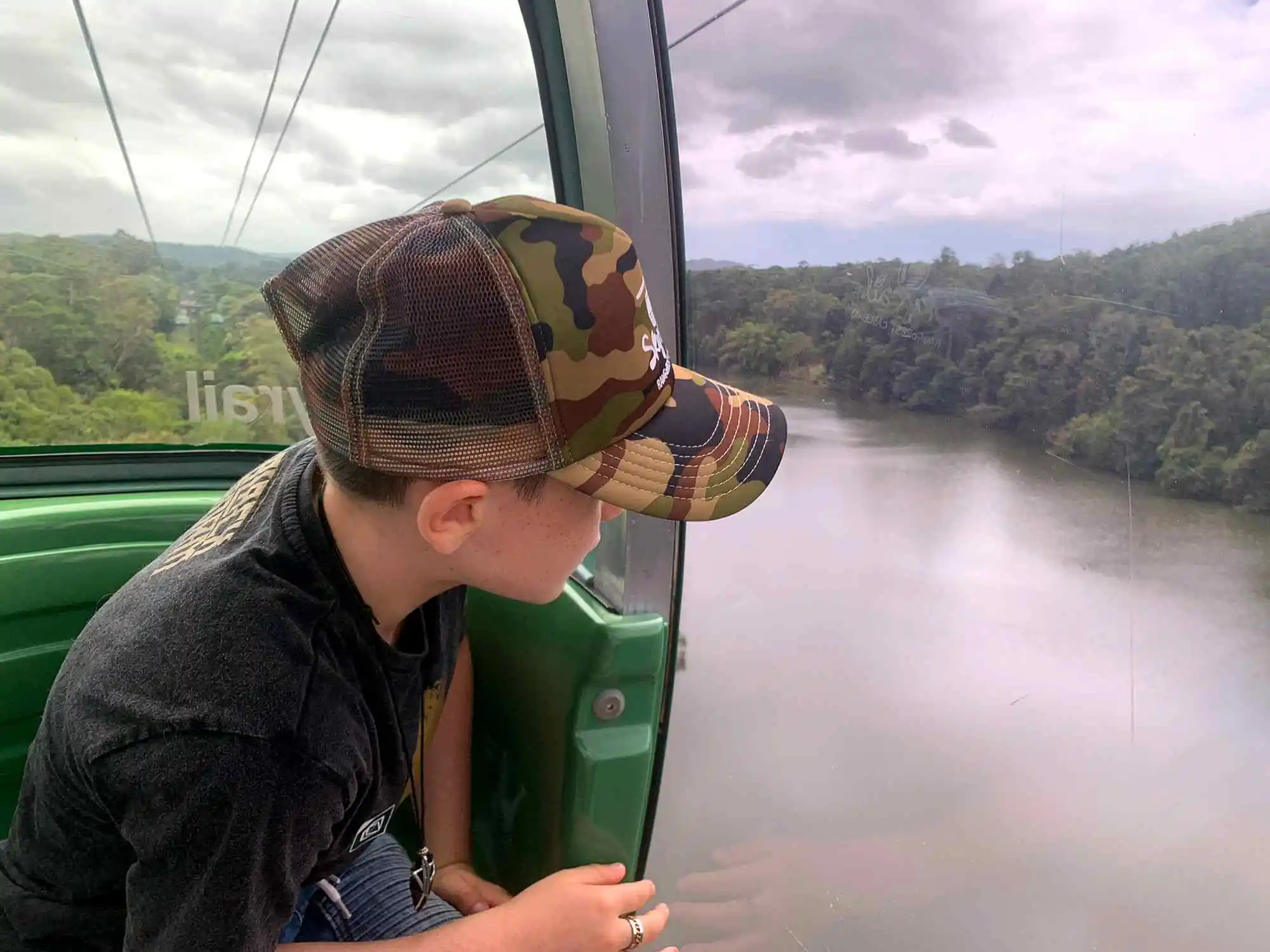 A side view of a boy wearing a cap, looking out of a gondola window at the Barron River below.