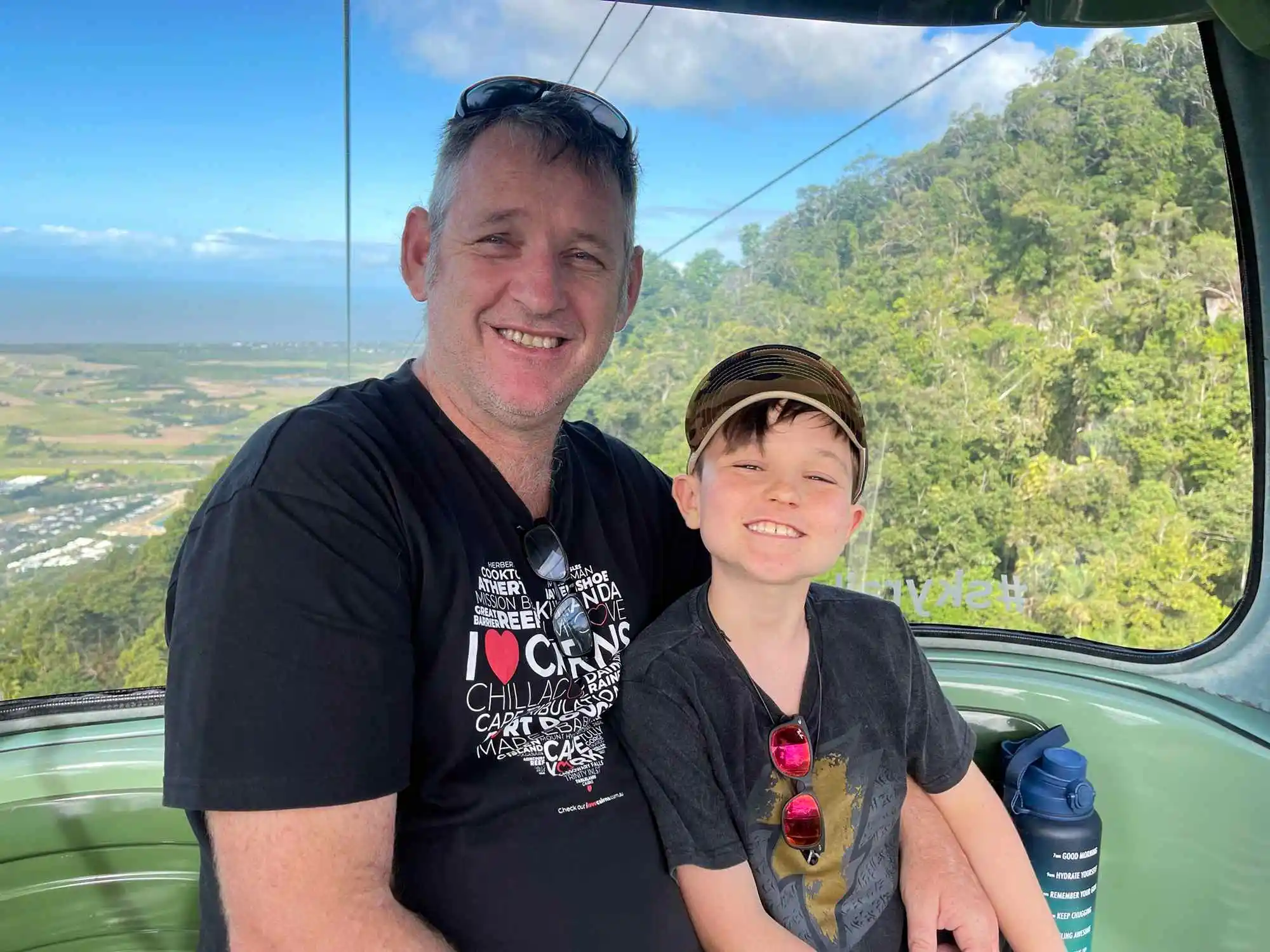 A man and boy smile at the camera sitting in a gondola with bright blue skies and green rainforest behind them.