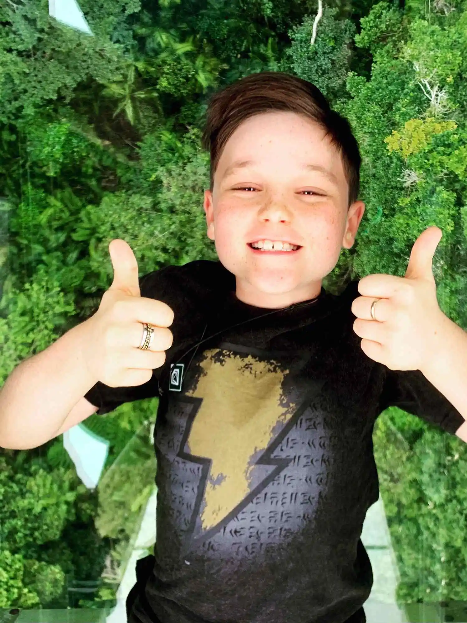 A boy lays on the glass floor with rainforest canopy below. Smiling and thumbs up t the camera.