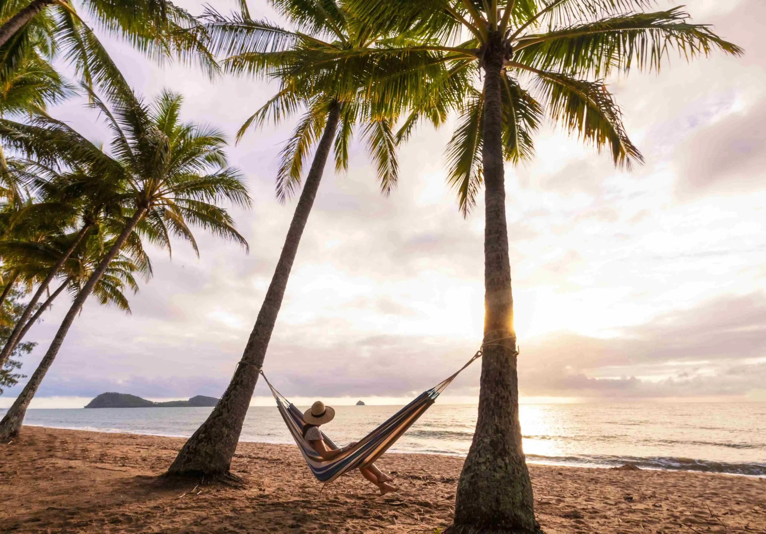 A girl in a large hat sits inside a hammock between two palm trees on the beach as the sun sets