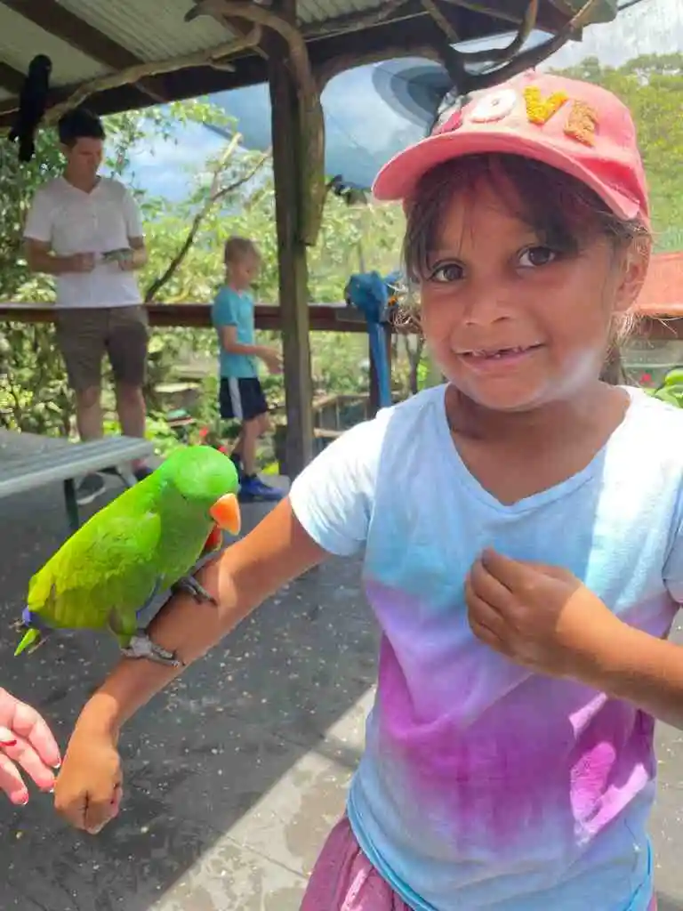 A green bird sits on a small girls arm as she smiles at the camera