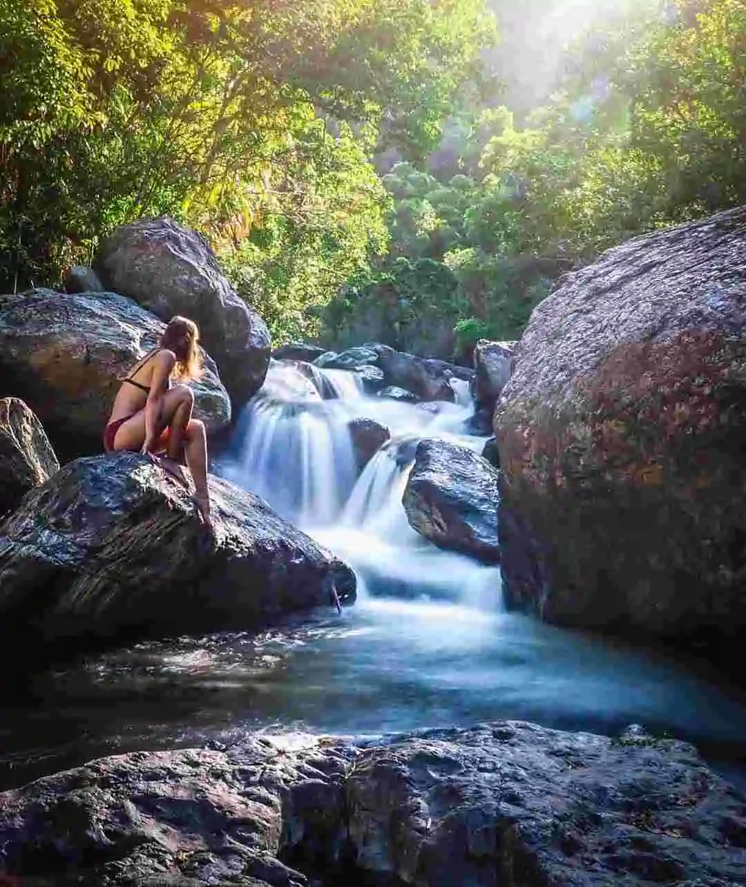 A woman sits in a swimsuit on a large rock looking at a waterfall as it cascades down the rocks