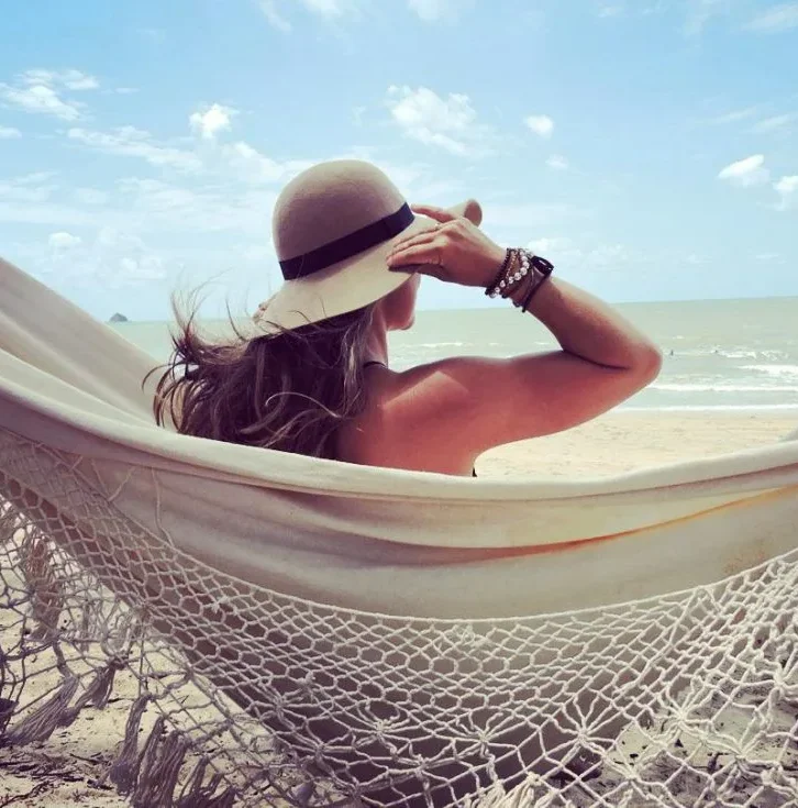 A woman sits in a hammock on a sandy beach holding onto her hat and looking out to sea