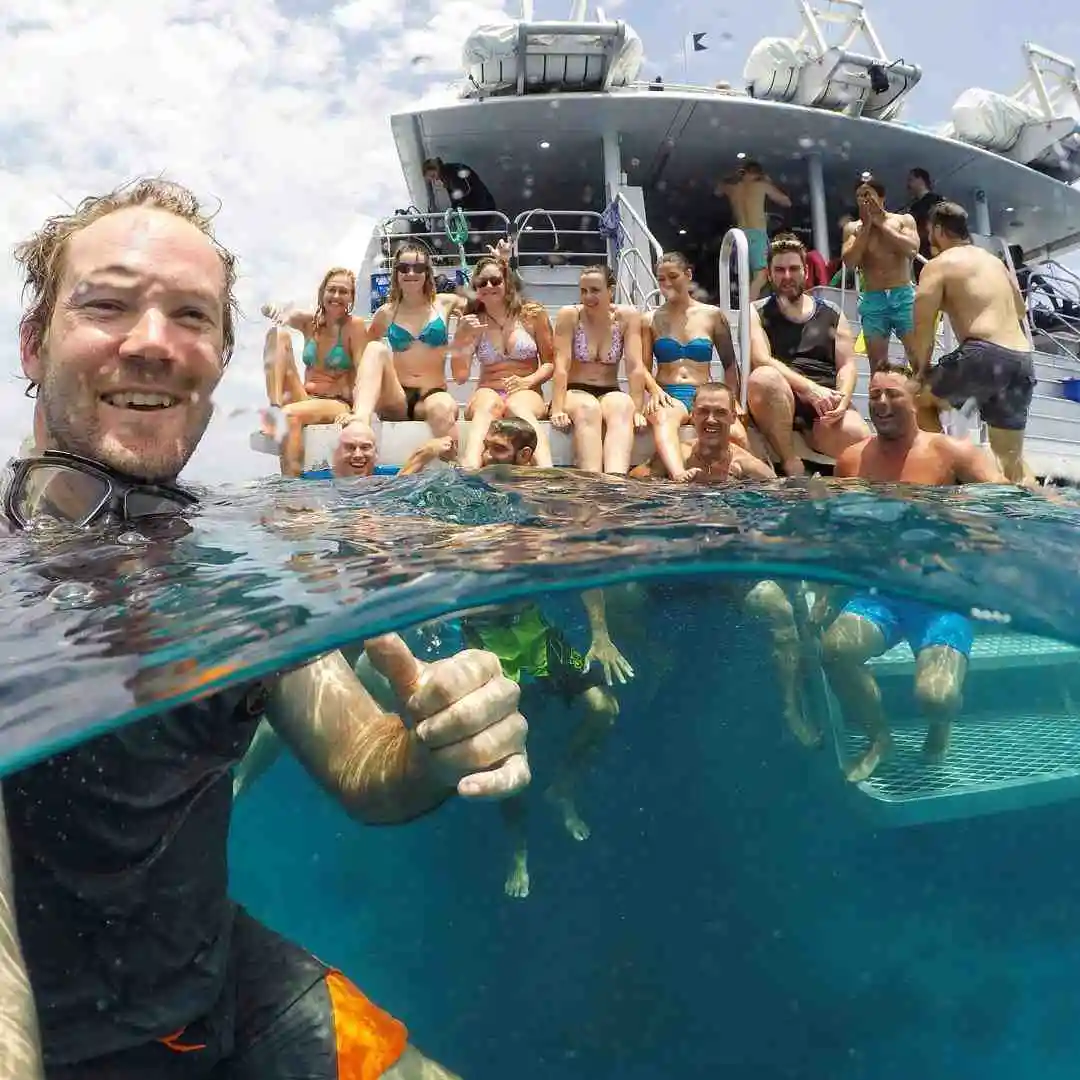 A man is in the forefront floating in the ocean with a thumbs up. A boat is behind him wiht a group of people with their arms up in the air. 