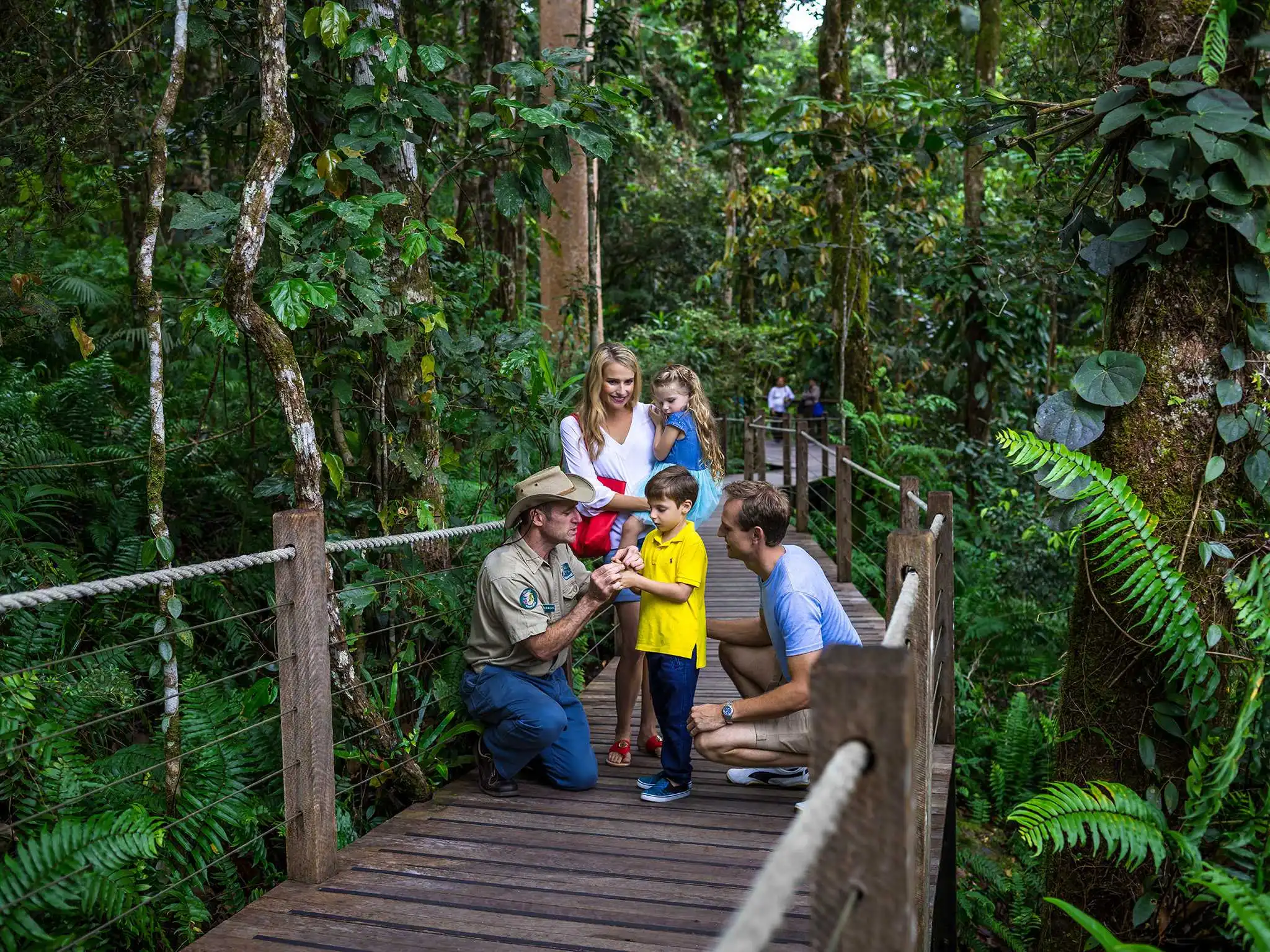 A ranger kneels on a rainforest boardwalk to talk to a small child and family