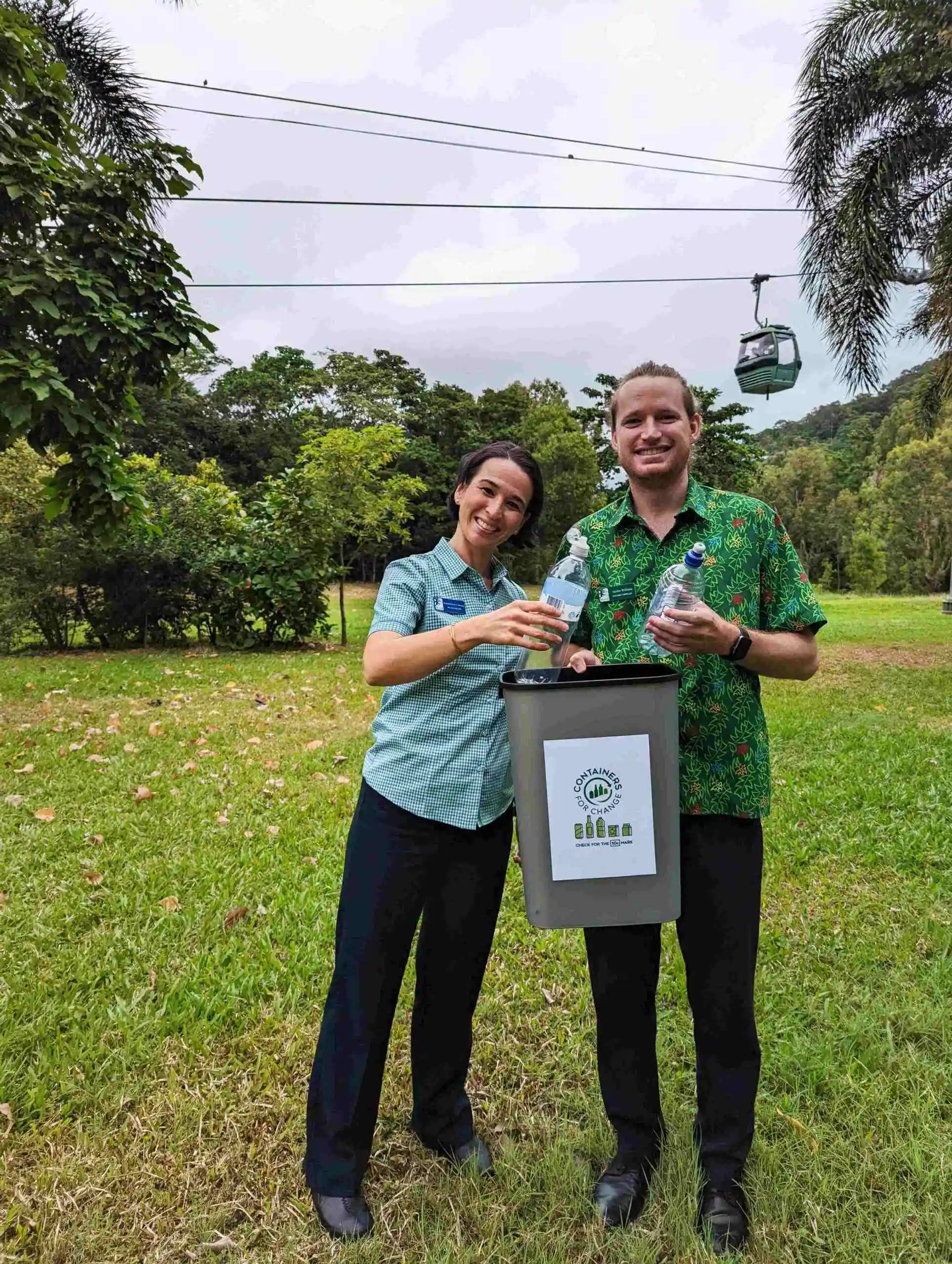 Two Skyrail staff with Containers for Change recycling contributing to a greener future standing on the lawn in uniform in front of the cableway