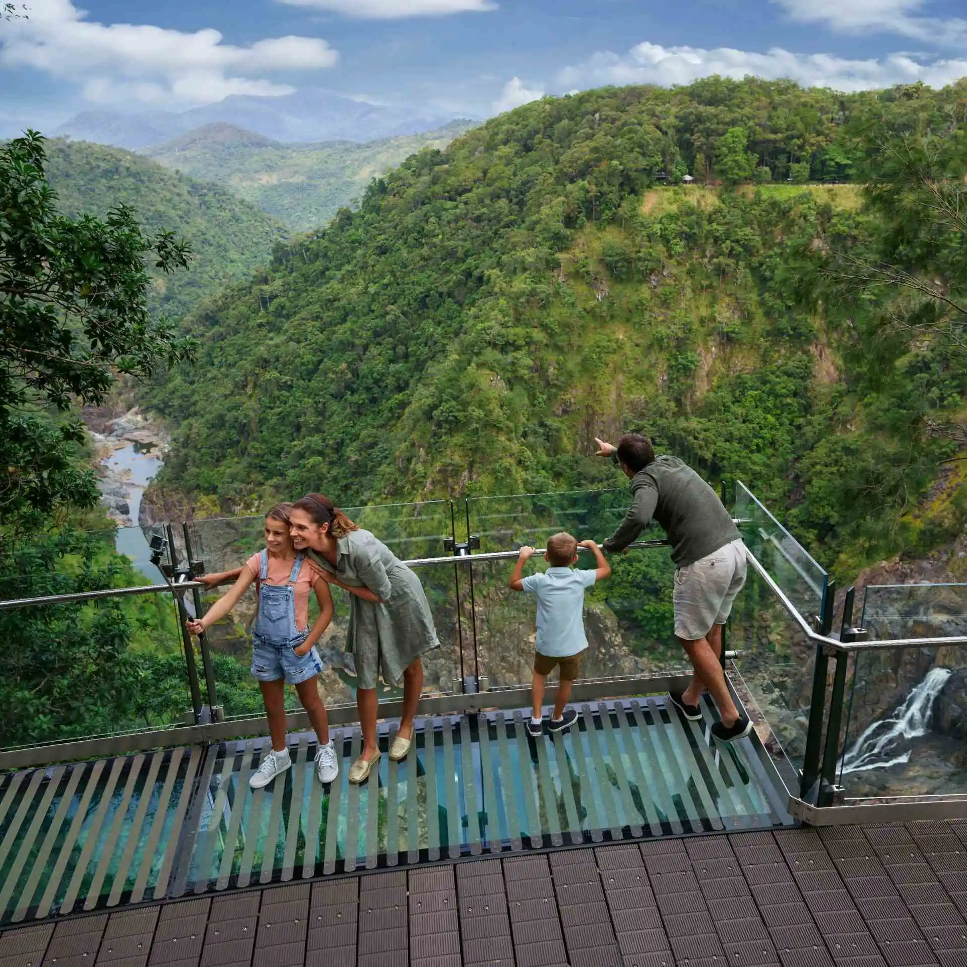 A woman and girls smile while taking a selfie. Behind them a  man and boy look out into the deep gorge as they stand on a glass walkway and edge