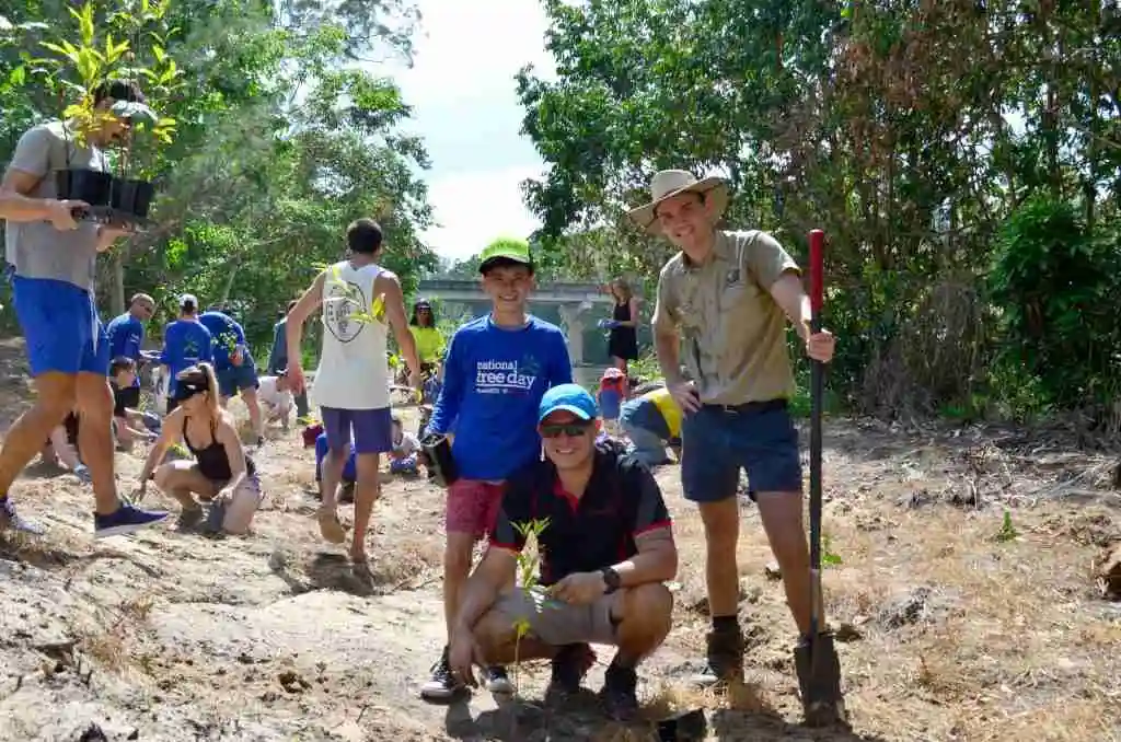 A man crouches on the ground with  a fresh tree planted. A Ranger and smal boy stand behind him, all smiling at the camera