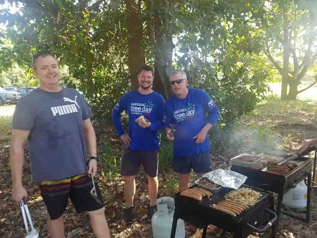Three men stand smiling in front of  a tree and next to a bbq cooking sausages and onions