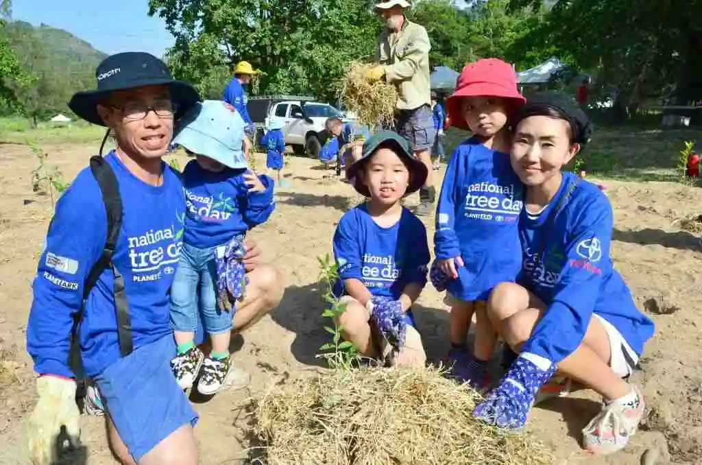 A male, femal and three small children all wearing blue hats and tree planting tee shirts kneel beside a freshly planted native tree