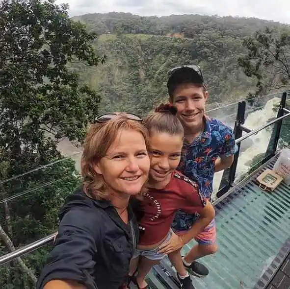 A woman and two children smile up at the camera on a glass lookout with a waterfall behind them.