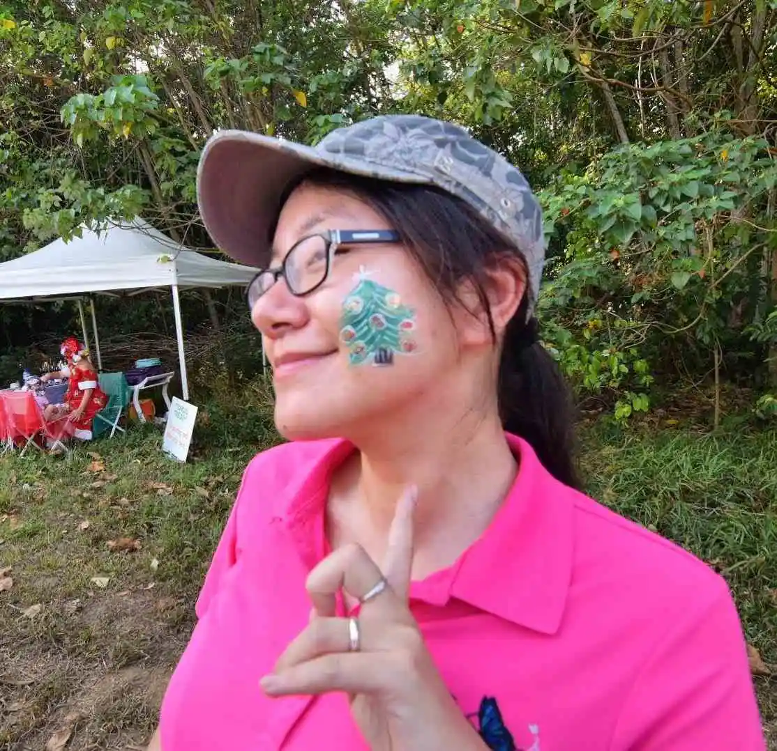 A girl in a pink shirt smiles and points to a glitter Christmas tree painted on her cheek