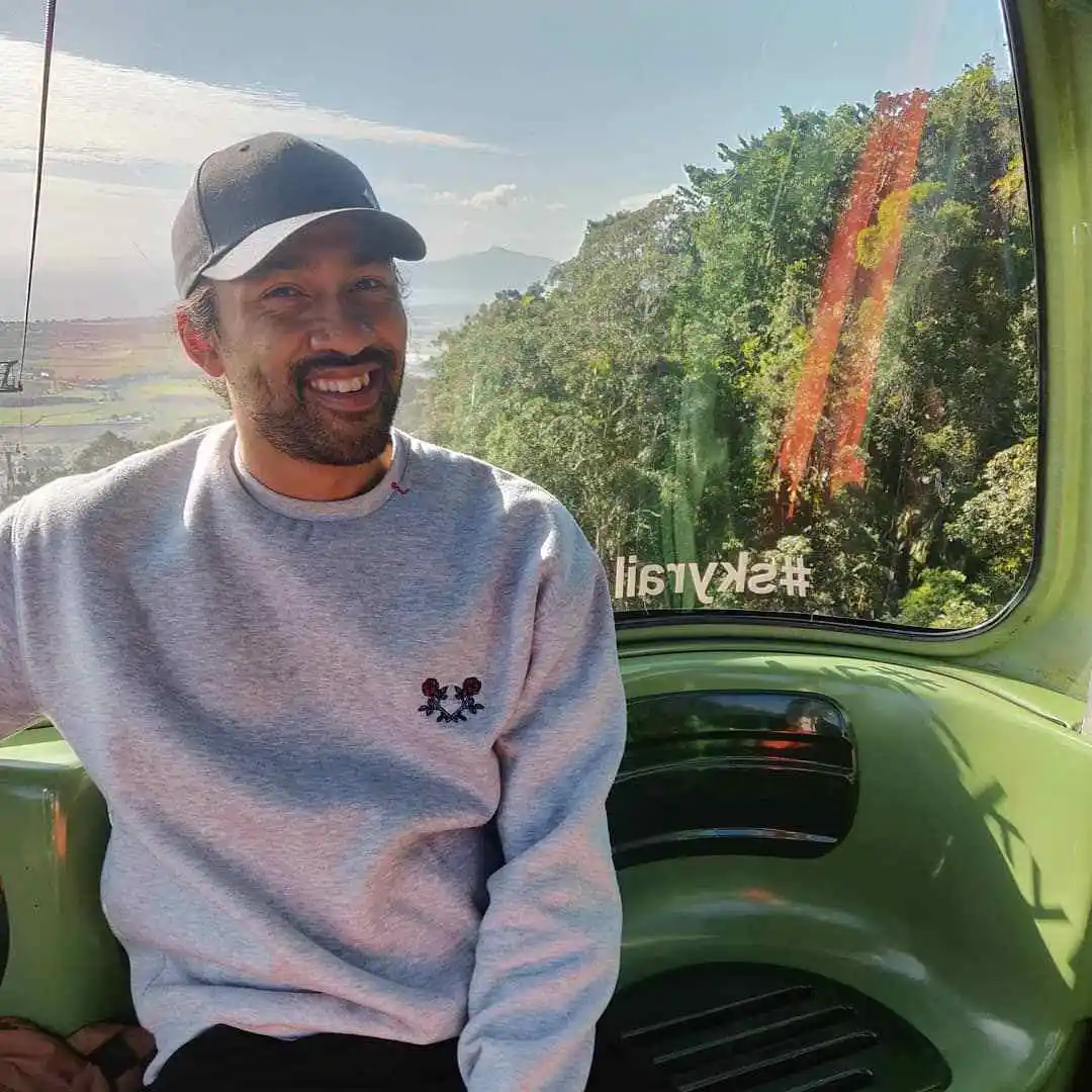 A man in a grey jumper sits in a gondola, smiling at the camera. Rainforest and blue skies behind him through the window.