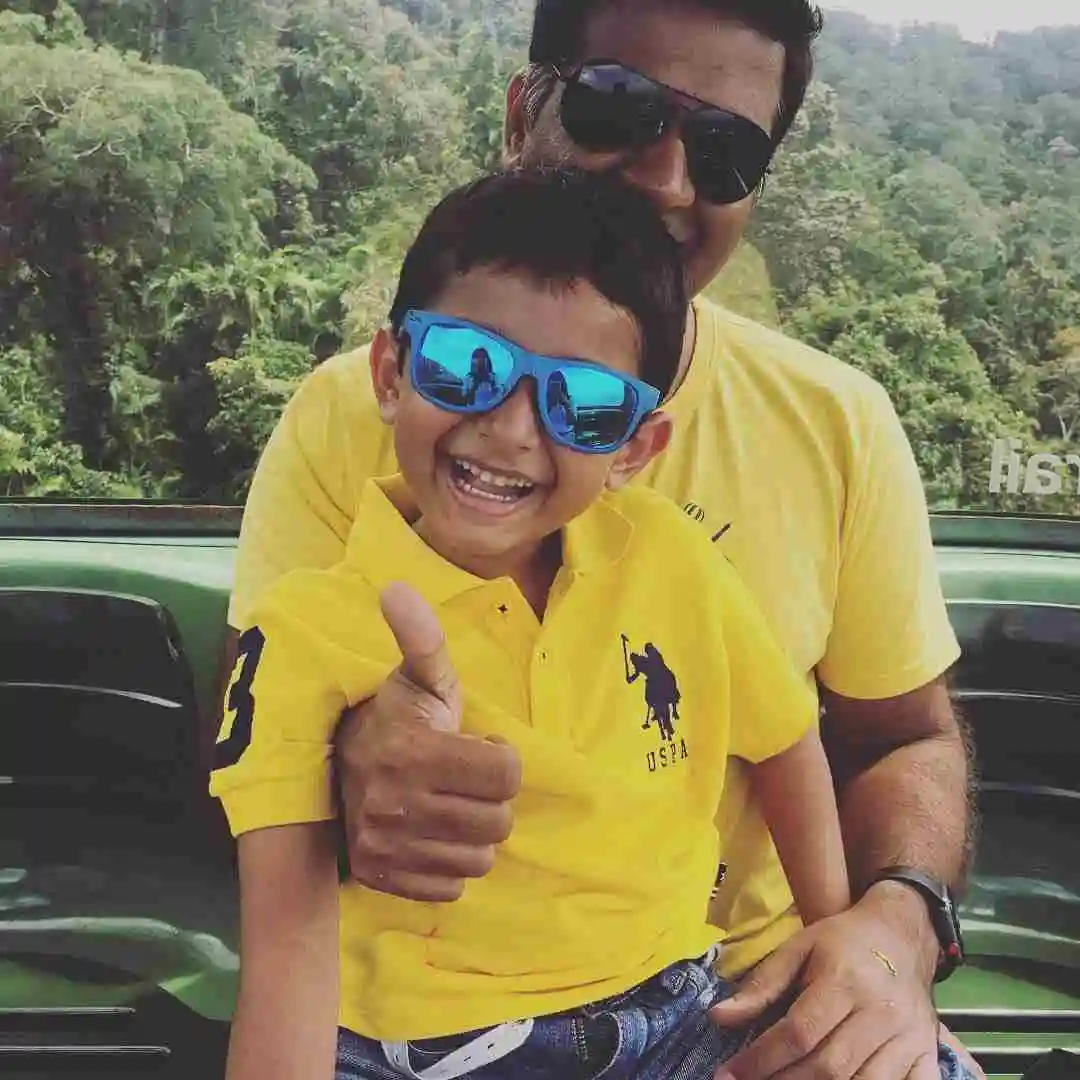 A boy in a yellow shirt sits on a mans knee. They are both smiling at the camera, the man has thumbs up