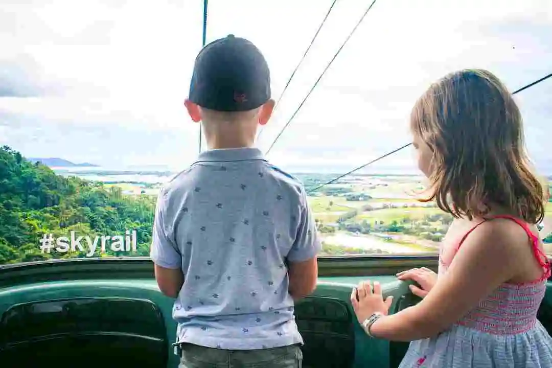 The back of two small children looking out of a gondola window into the rainforest below and blue skies