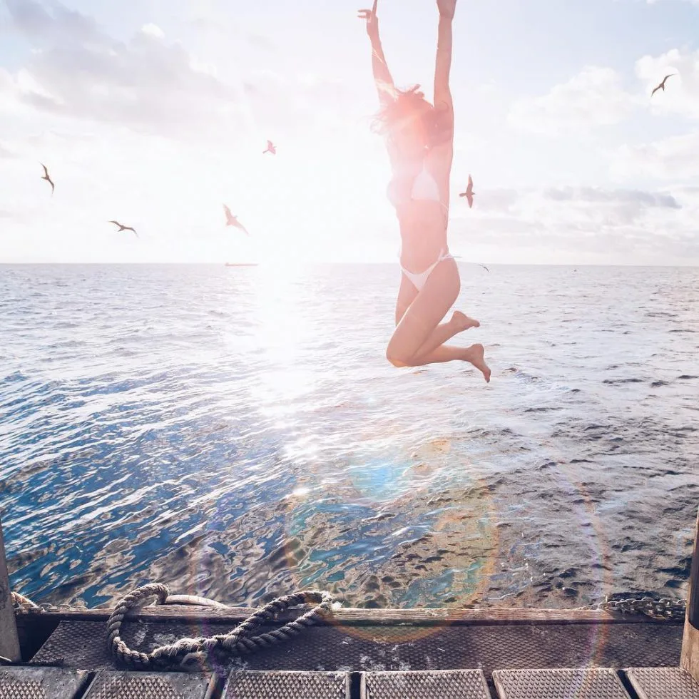 A woman jumps in the air on a wooden boardwalk next to the ocean with the sun glaring in the background