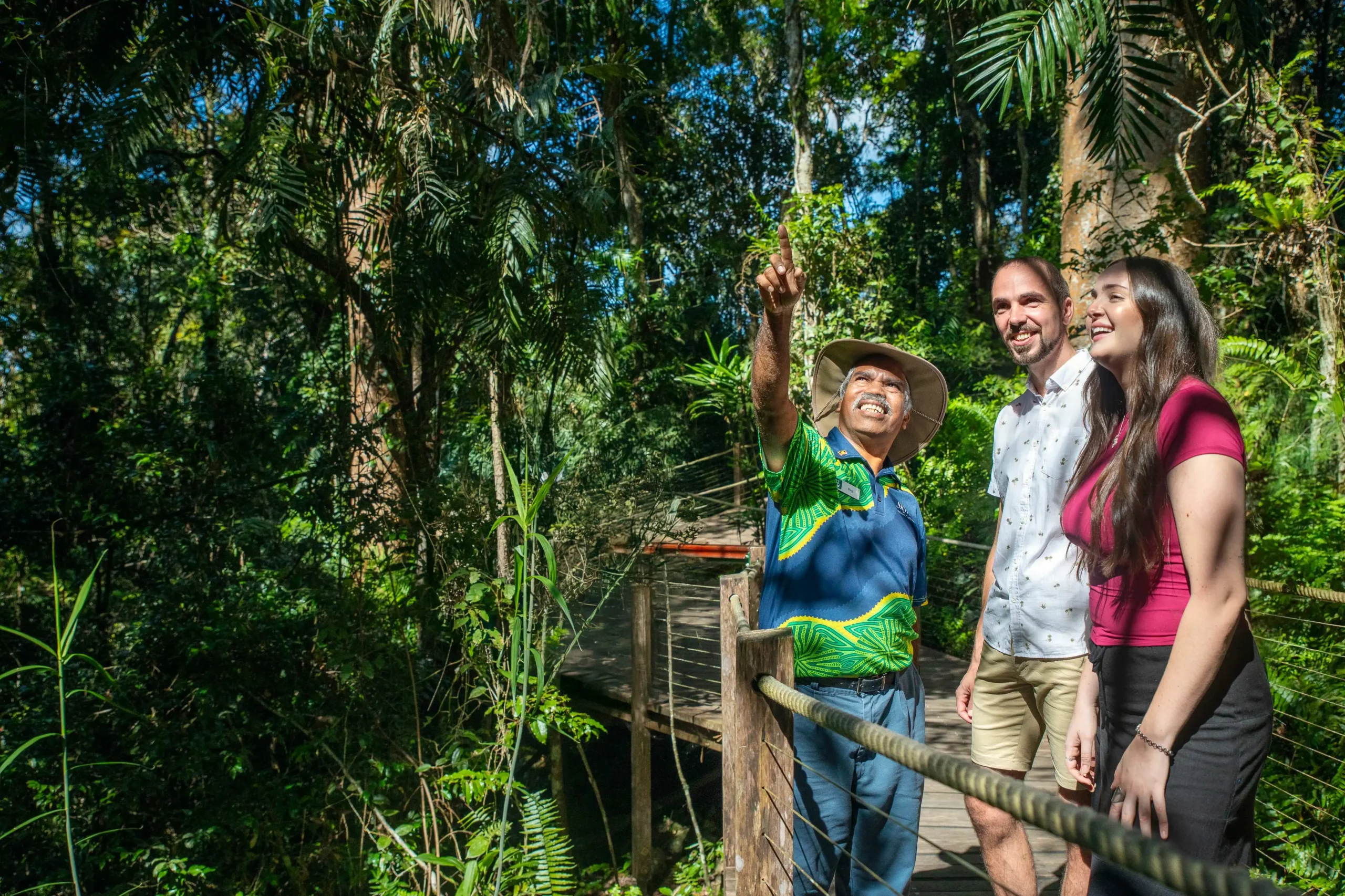 Two people stand on a boardwalk surrounded by tall trees, looking up at the sky as a Ranger points into the canopy. 