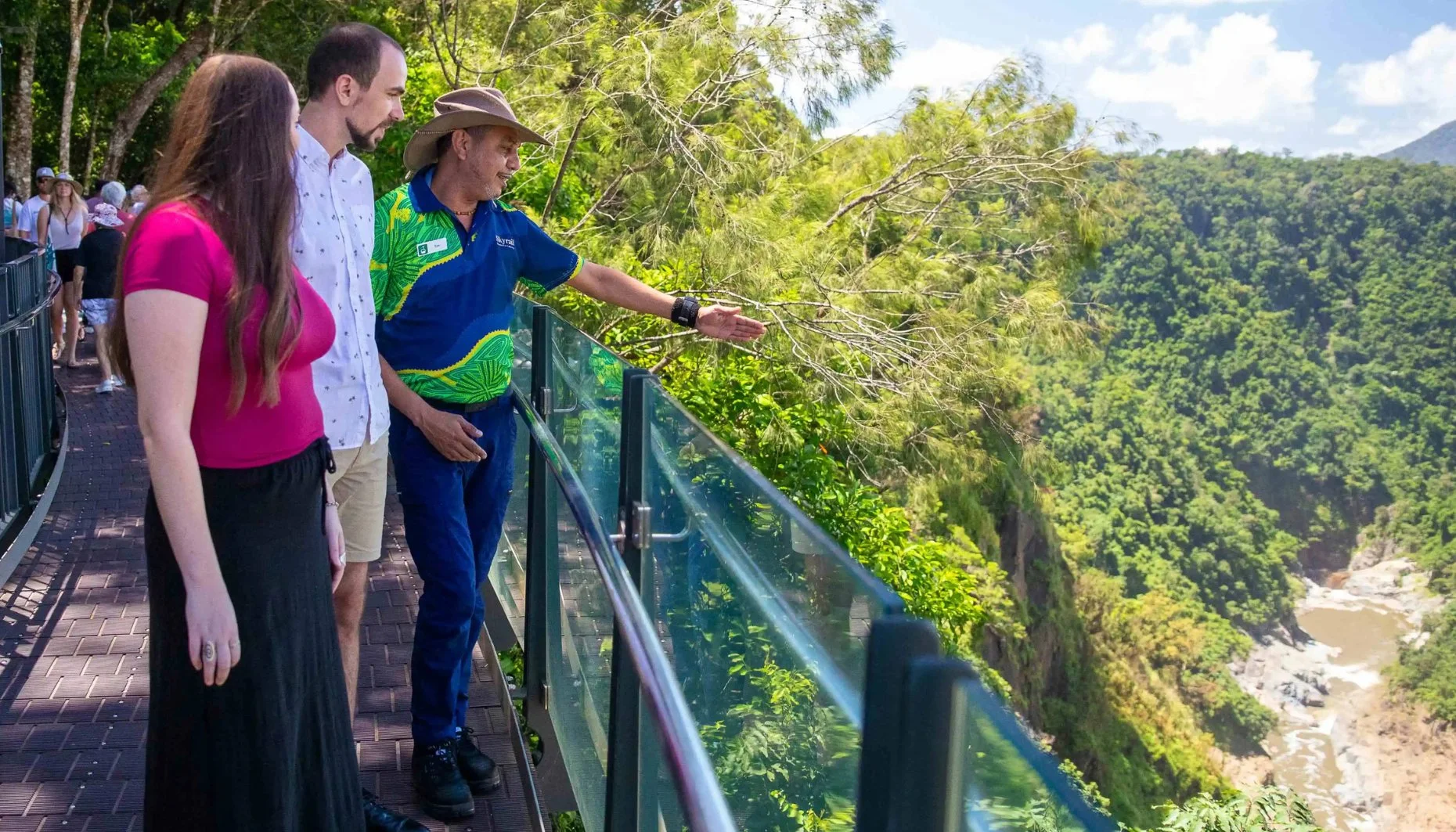 A Ranger and two people stand on a lookout, the Ranger is pointing into the distance into the deep gorge.
