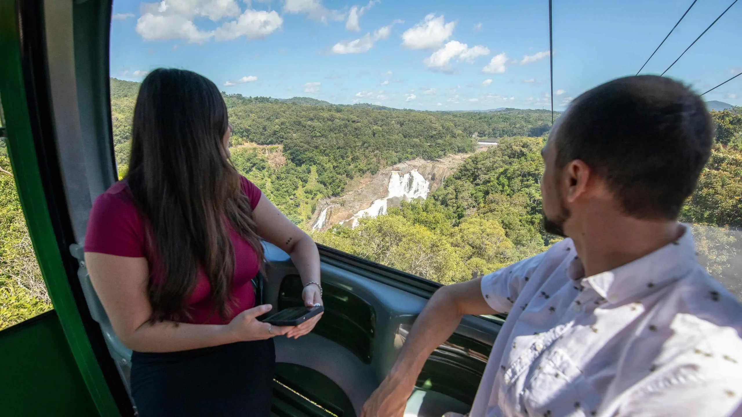 Two people sit turned away looking out of a gondola window at a rocky waterfalls in the rainforest