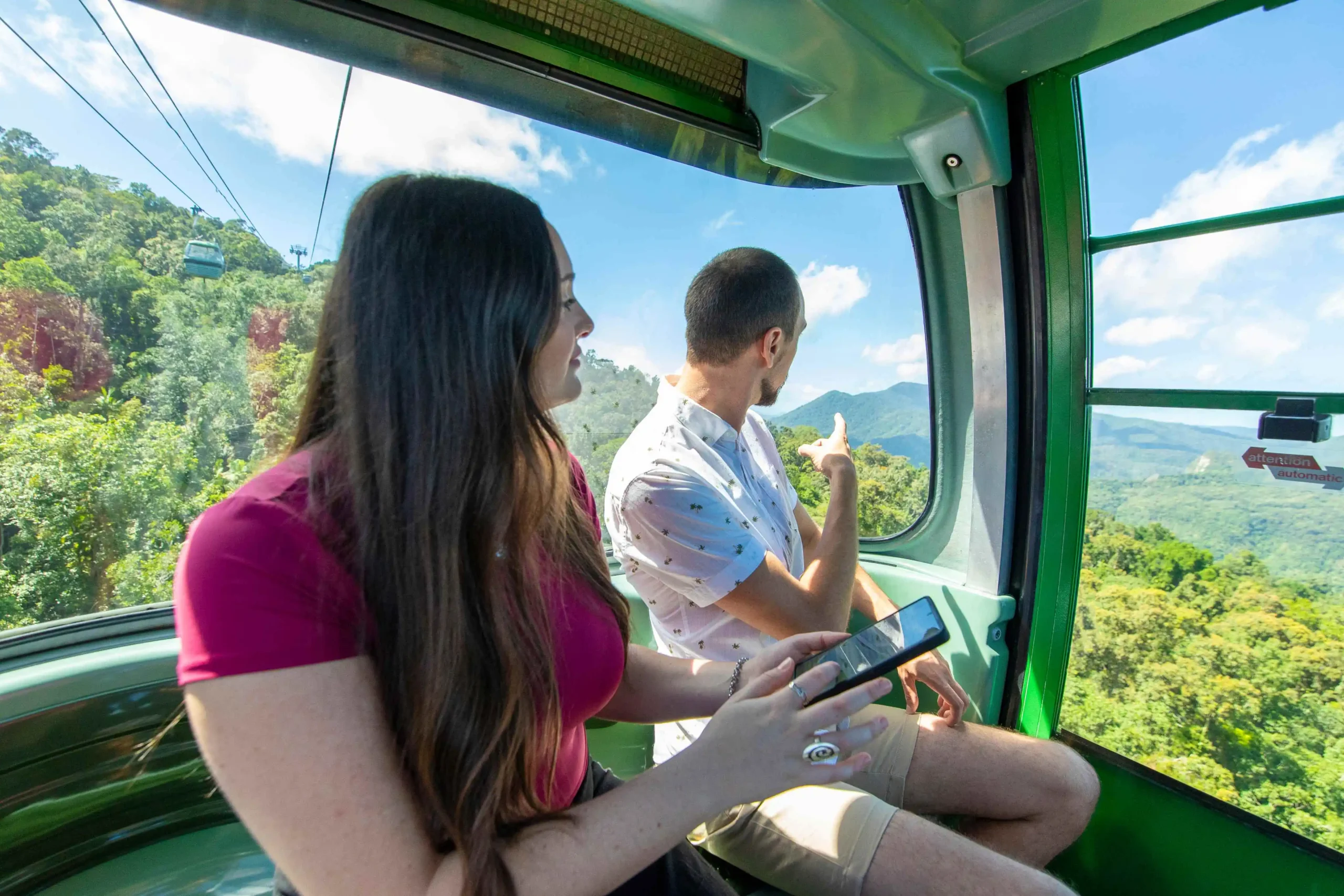 Two people sit holding a phone, turned looking out of a gondola window at the blue skies and lush green rainforest below. 